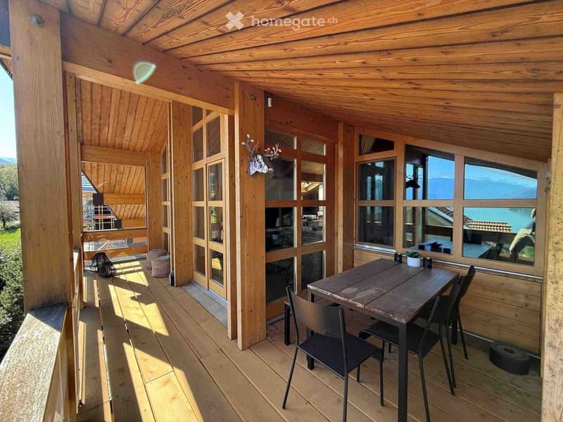 Wooden balcony with large windows offering a scenic view, featuring a table, chairs, a bench, and a small potted plant.
