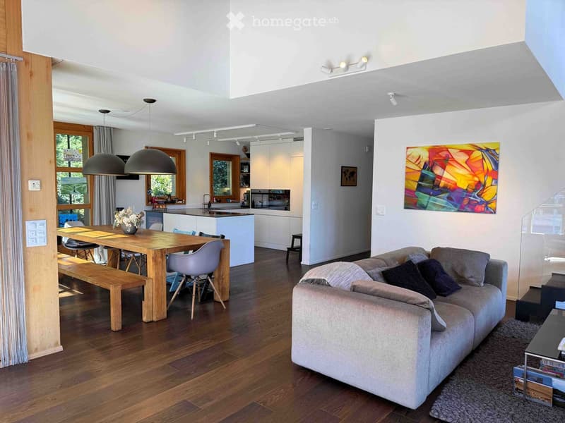 Open-concept living and dining area with a grey sectional sofa, coffee table, wooden dining table with chairs, pendant lights, and a modern kitchen in the background.