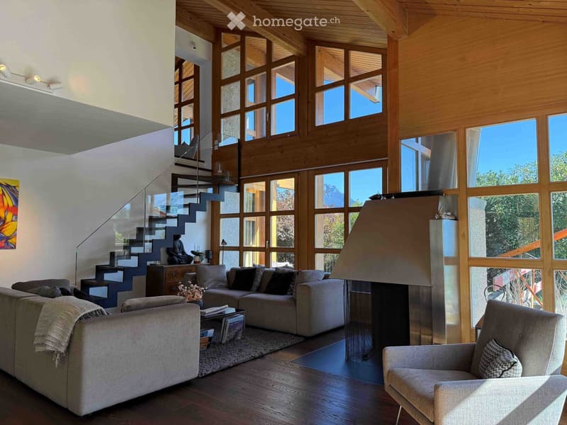 Modern living room with large windows, wooden ceiling, and a view. Features a grey sectional sofa, coffee table, armchair, fireplace, and a staircase leading to an upper level.
