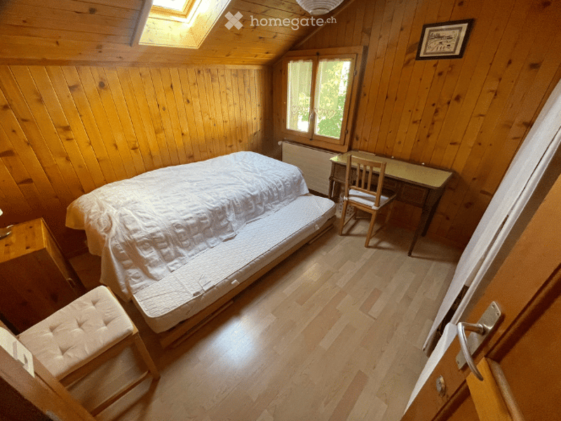 Cozy attic bedroom with wooden walls and floors, featuring a single bed, a small desk with a chair, and a cushioned stool near the bed.