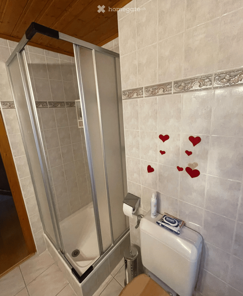 Bathroom with a glass-enclosed shower, toilet, toilet paper holder, a small bottle on the toilet tank, and a wooden stool in front of the toilet. The walls are tiled with decorative borders and small red heart stickers.