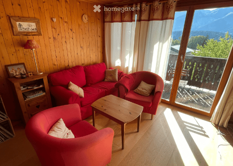 Cozy living room with wooden walls and floors, red sofa, armchairs, coffee table, and a view of mountains through large windows.