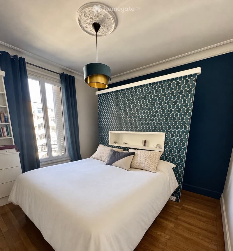 Modern bedroom with a large bed, decorative pillows, a built-in shelf, a hanging lamp, and a window with dark curtains.