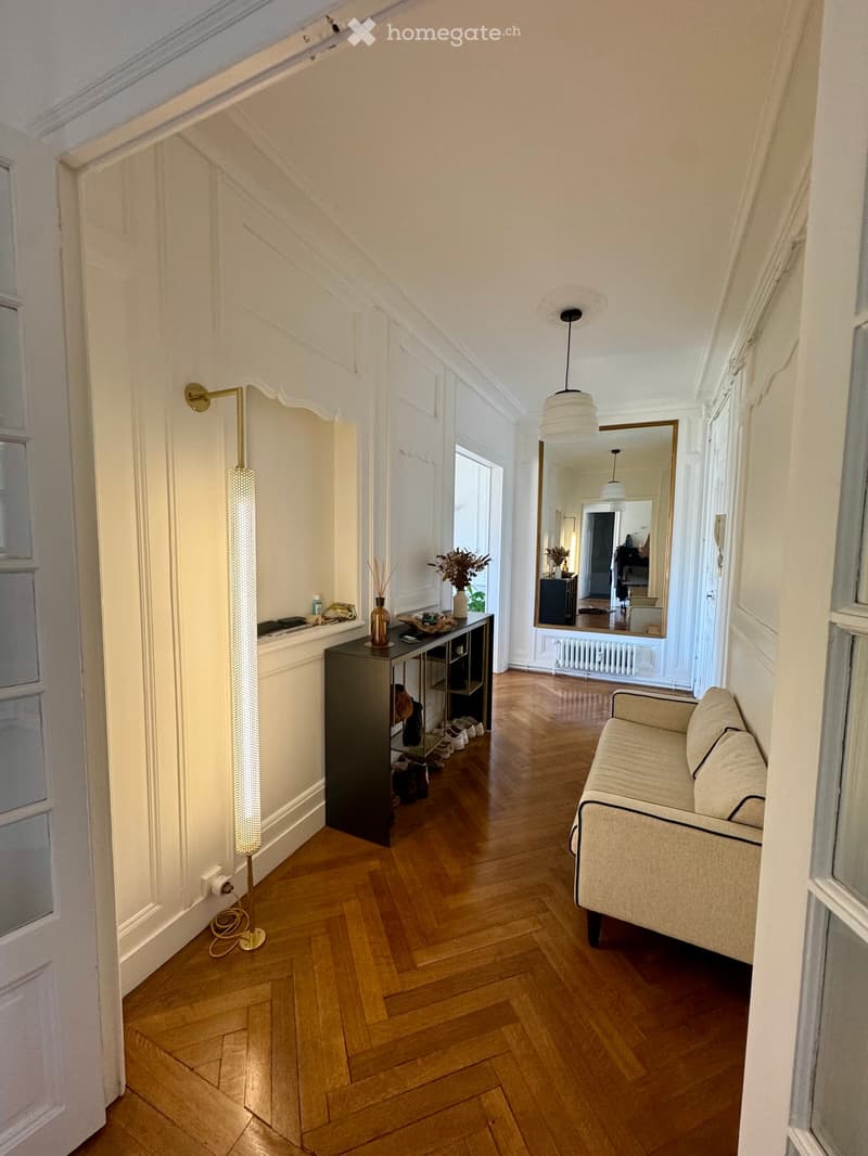 A well-lit hallway with wooden flooring, a sofa on the right, a small table with a vase and decorative items, a tall floor lamp, and a large mirror reflecting part of the room.