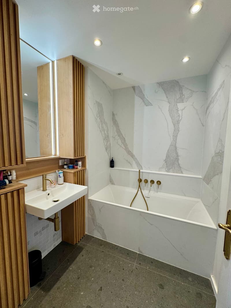 Modern bathroom with a freestanding bathtub, marble walls, a white sink with a gold faucet, and wooden cabinets.