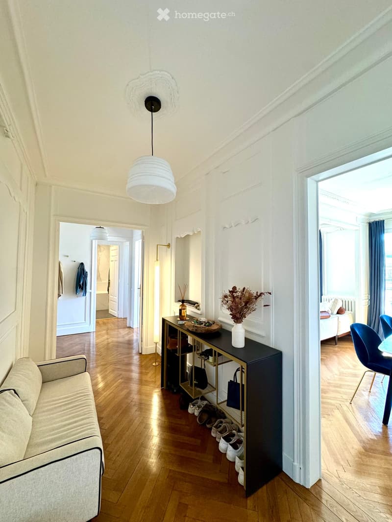 A modern hallway with wooden flooring, a sofa on the left, a small table with a vase and decorative items, and a shelf holding shoes and bags. A pendant light hangs from the ceiling, and a doorway leads to another room.