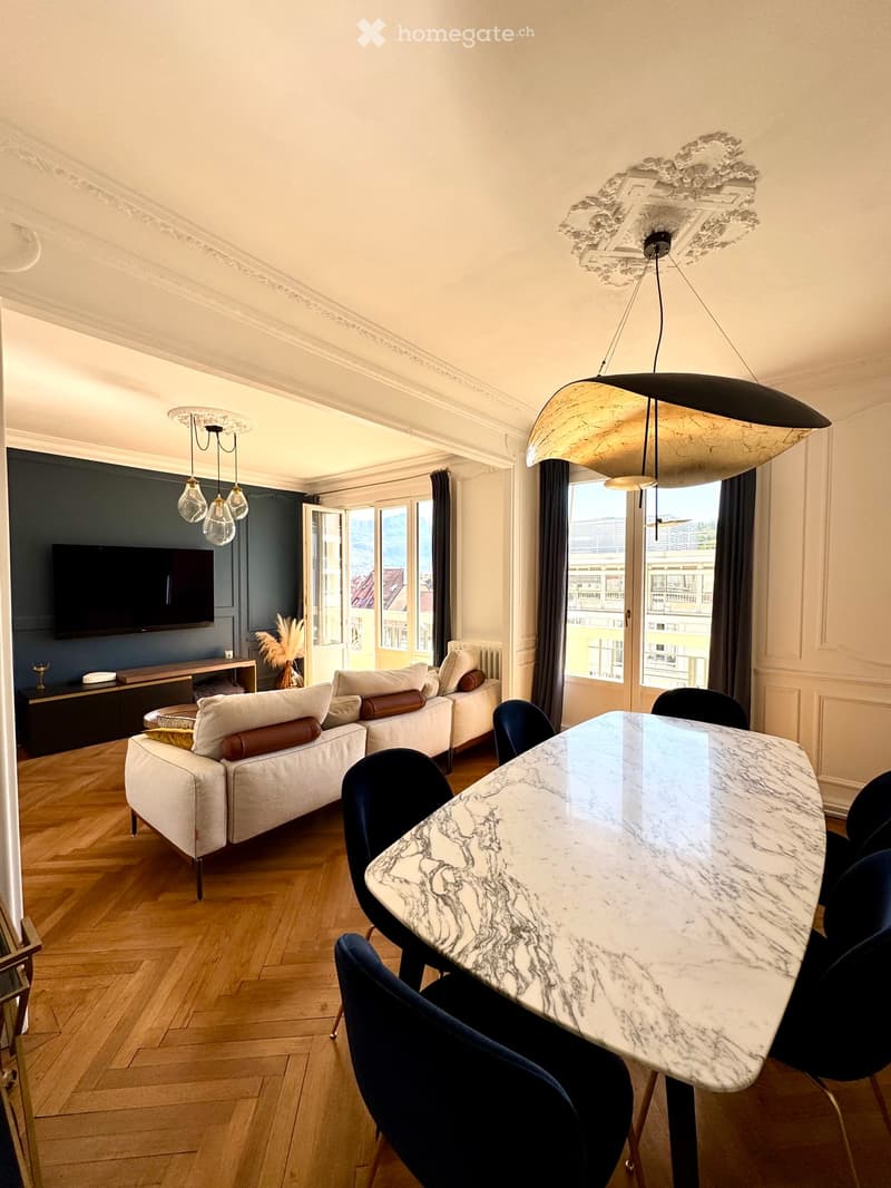 Modern living room with a white sofa, marble dining table, dark blue chairs, large windows with curtains, and a television on a wooden stand.