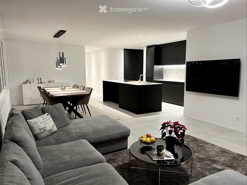 Modern living and dining area with a grey sectional sofa, coffee table, dining table with chairs, and a kitchen island with black cabinets and a stainless steel sink.