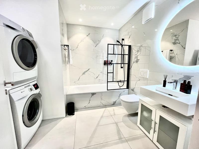 Modern bathroom with white marble tiles, a large sink, toilet, bathtub, washing machine, and a sleek mirror.