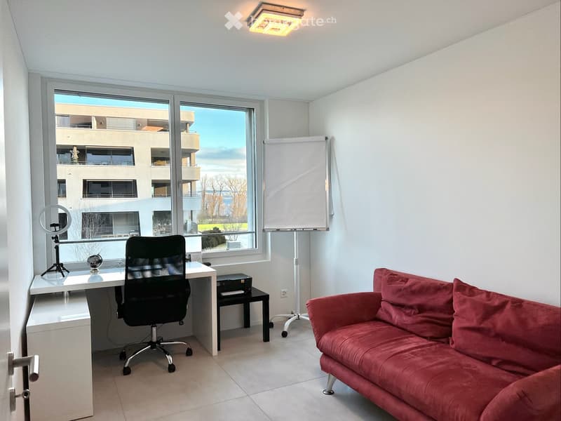 Modern furnished room with a red sofa, white desk, black chair, and large windows offering a view of the building outside.