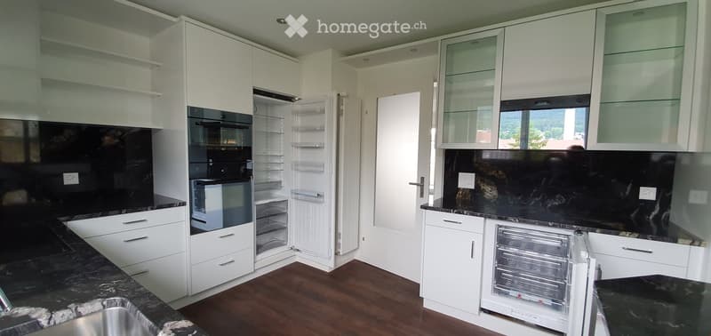 Modern kitchen with white cabinets, black countertops, and stainless steel appliances including a refrigerator and oven. The floor is wooden, and the space is well-lit with natural light from a window.