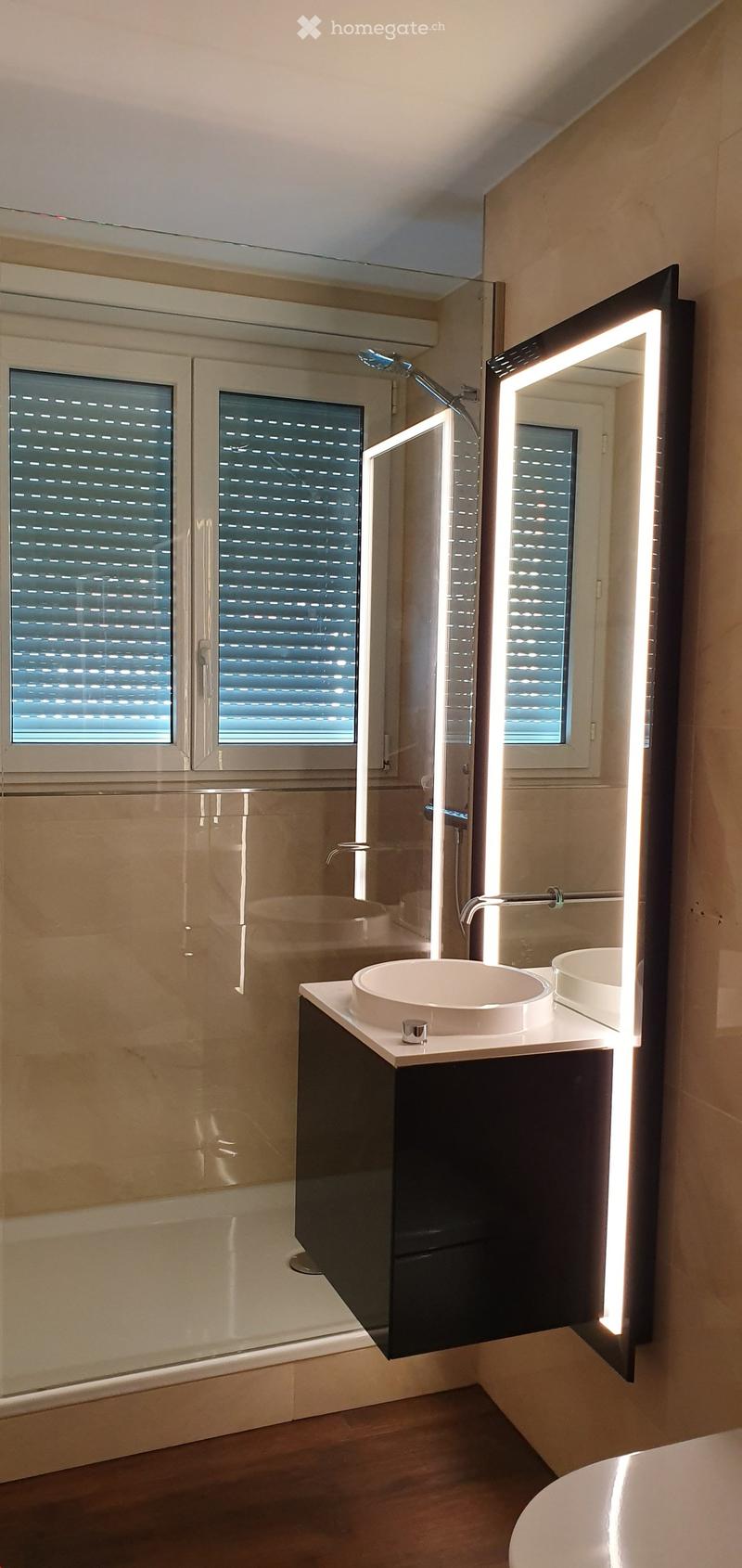 Modern bathroom with a sleek black vanity, two white sinks, a large illuminated mirror, and a glass-enclosed shower area with a modern showerhead.