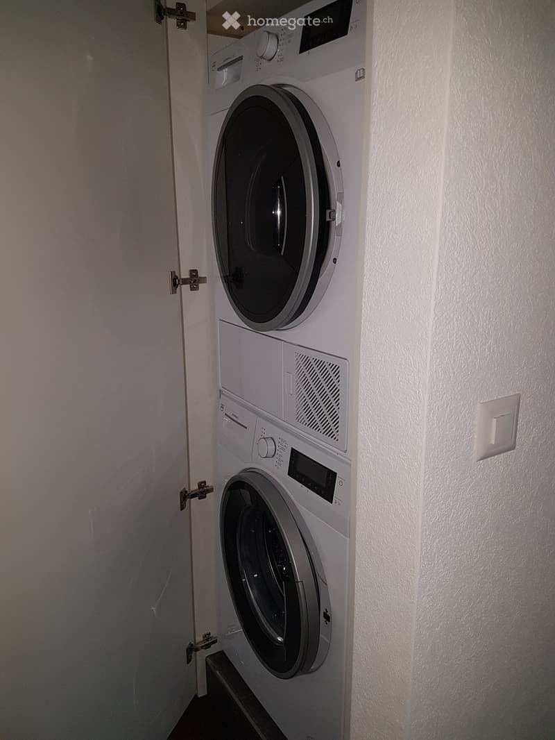 Compact space with a stacked washing machine and dryer unit, white walls, and a door with a latch.