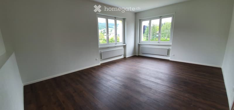 Empty room with wooden flooring, light grey walls, and large windows offering a view of greenery outside. Radiators are visible under the windows.