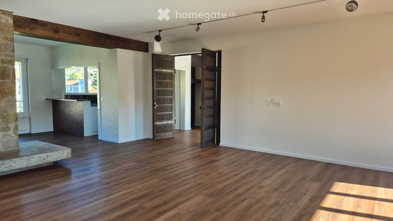 Empty room with wooden flooring, white walls, and a stone pillar. There is an open doorway leading to another room with a kitchen area visible in the background.