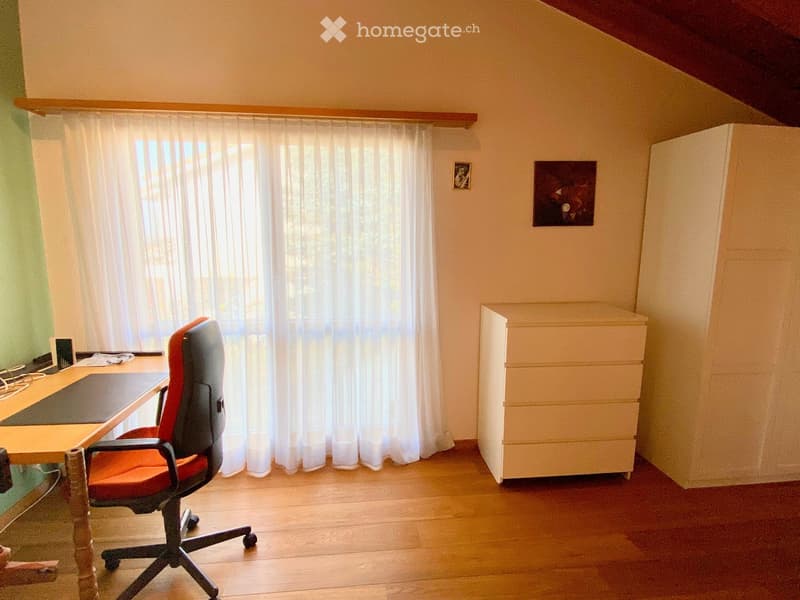 A small, well-lit room with wooden flooring, a large window with white curtains, a red office chair, a wooden desk, and a white cabinet.