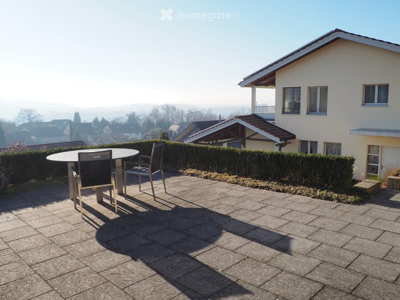 Paved patio with a round table and two chairs, surrounded by hedges and a view of a residential area.