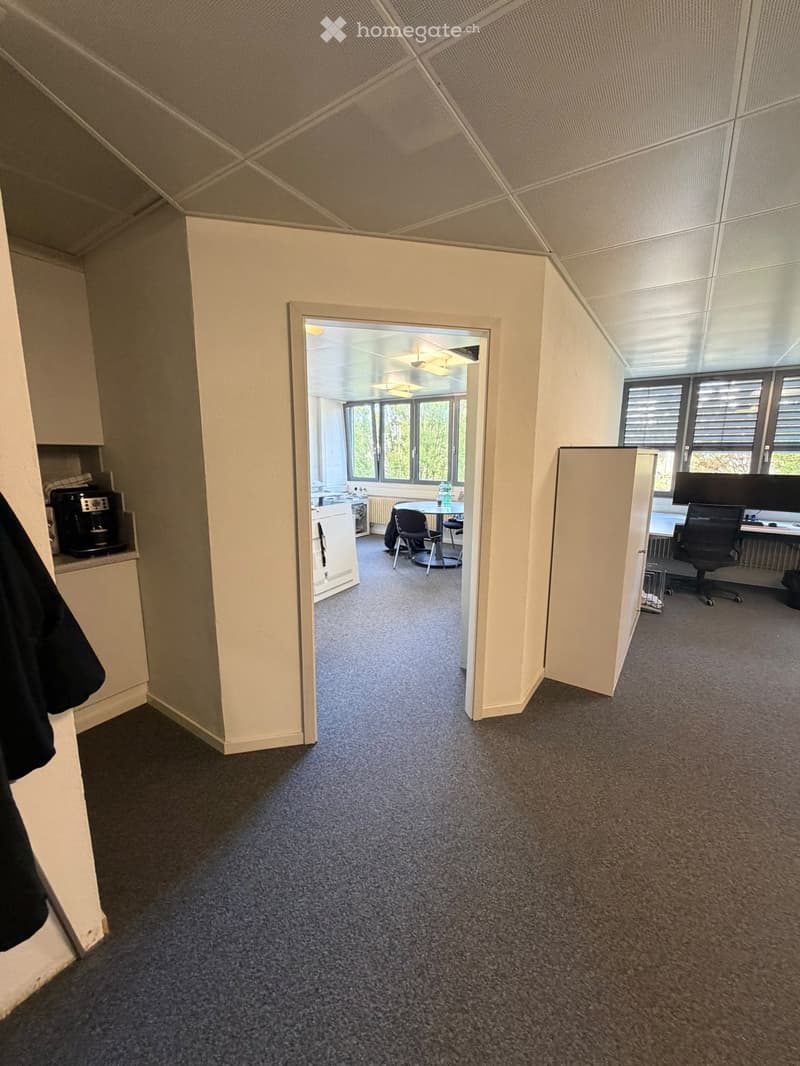 Office space with carpeted floor, white walls, and a slanted ceiling. Visible objects include a chair, table, screen, cupboard, and coffee machine.