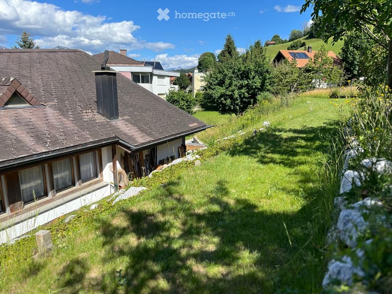A two-story house with a dark shingled roof, white walls, and multiple windows, situated on a grassy slope with surrounding trees and hills in the background.