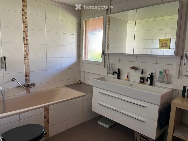 Modern bathroom with a bathtub, double sink, large mirror, and a toilet. The walls are tiled with a decorative mosaic border, and the floor is tiled in a warm brown color. A small window provides natural light.