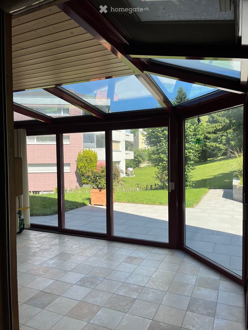Bright, spacious sunroom with large glass sliding doors opening to a tiled patio and lush garden. The room features a wooden slanted ceiling and ample natural light.