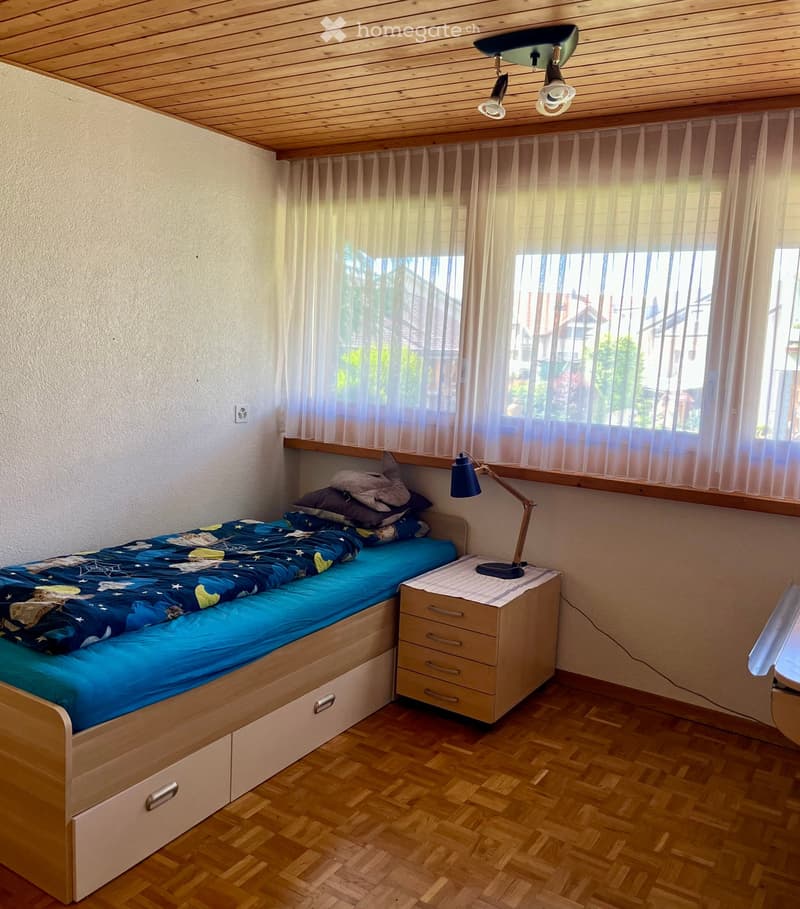 Cozy bedroom with a single bed, nightstand, and lamp. The bed has a blue duvet and pillows, and the room features wooden flooring and large windows with sheer curtains.