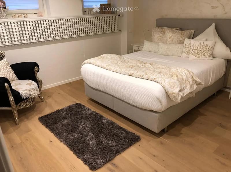 Cozy bedroom with a large bed, patterned pillows, a decorative throw, a plush grey rug, and a stylish black and gold armchair.