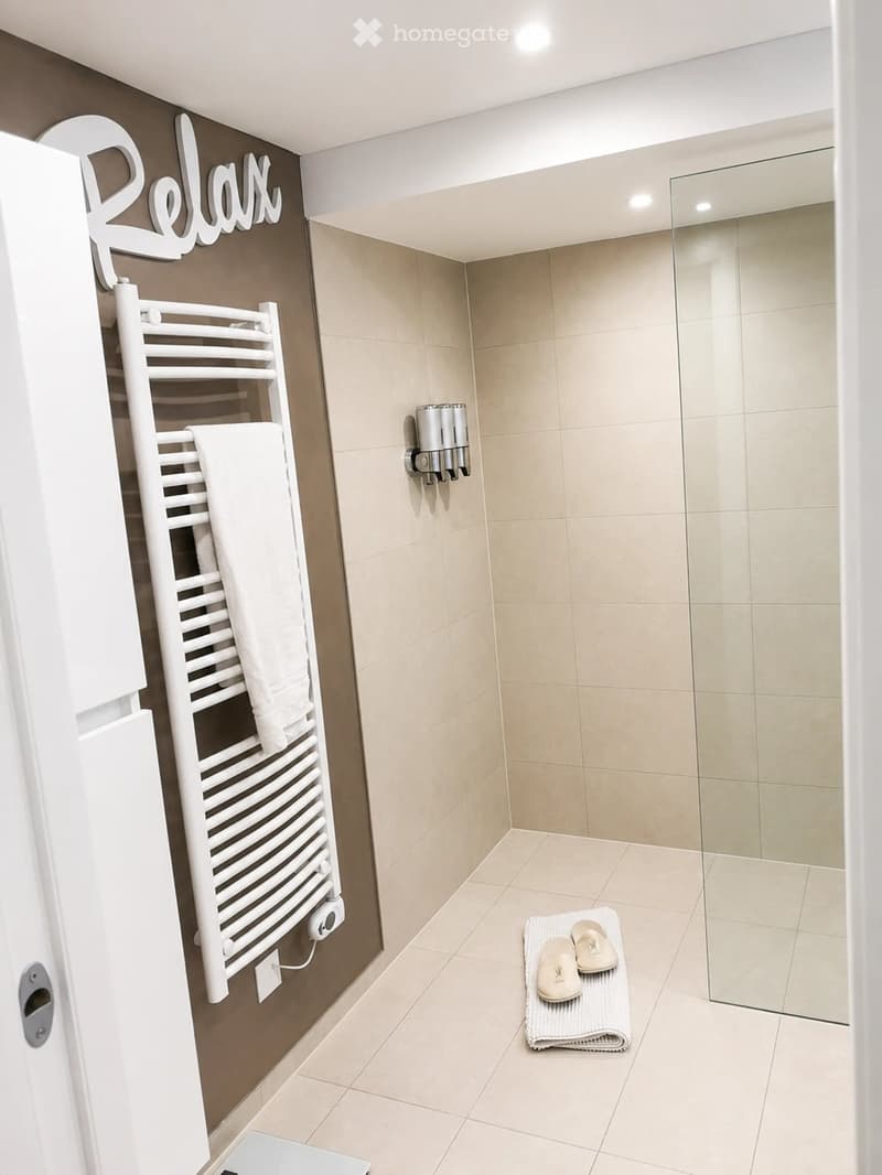 Modern bathroom with beige tiles, a white towel rack with towels, a bathroom scale, a soap dispenser, and a pair of slippers on a towel mat.