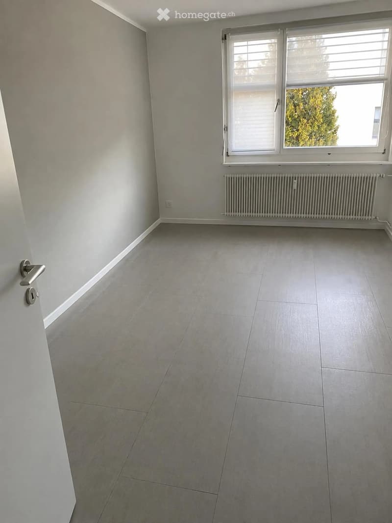 Empty room with light grey tiled floor, white walls, a window with blinds, and a radiator below it. The door handle is visible on the left side.