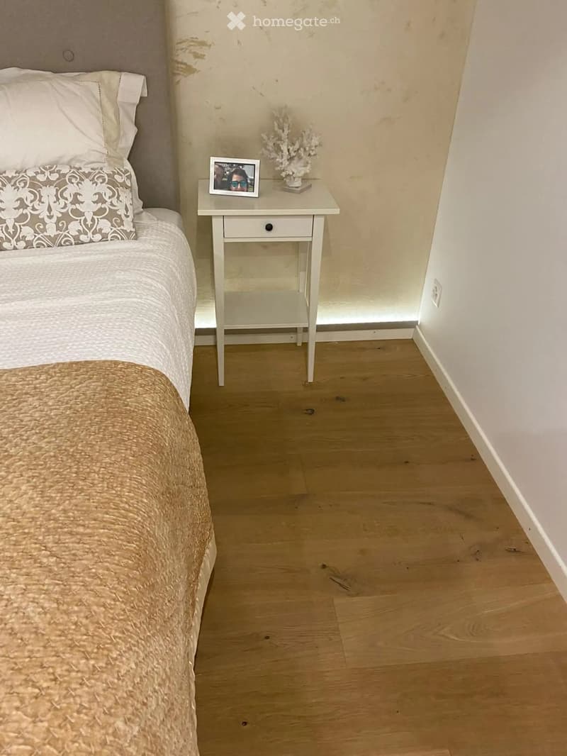 Cozy bedroom with a neatly made bed, a nightstand with a picture frame and decorative item, and light wooden flooring.