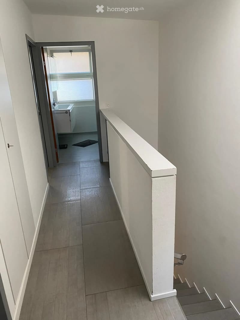 Empty hallway with tiled floor, white walls, and a staircase leading upwards. An open door leads to a bathroom with a sink, toilet, and bathtub.
