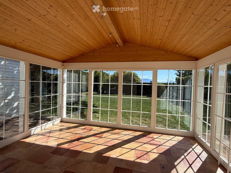 Bright sunroom with wooden ceiling, large glass windows, and terracotta tile flooring, offering a view of a green lawn and distant mountains.