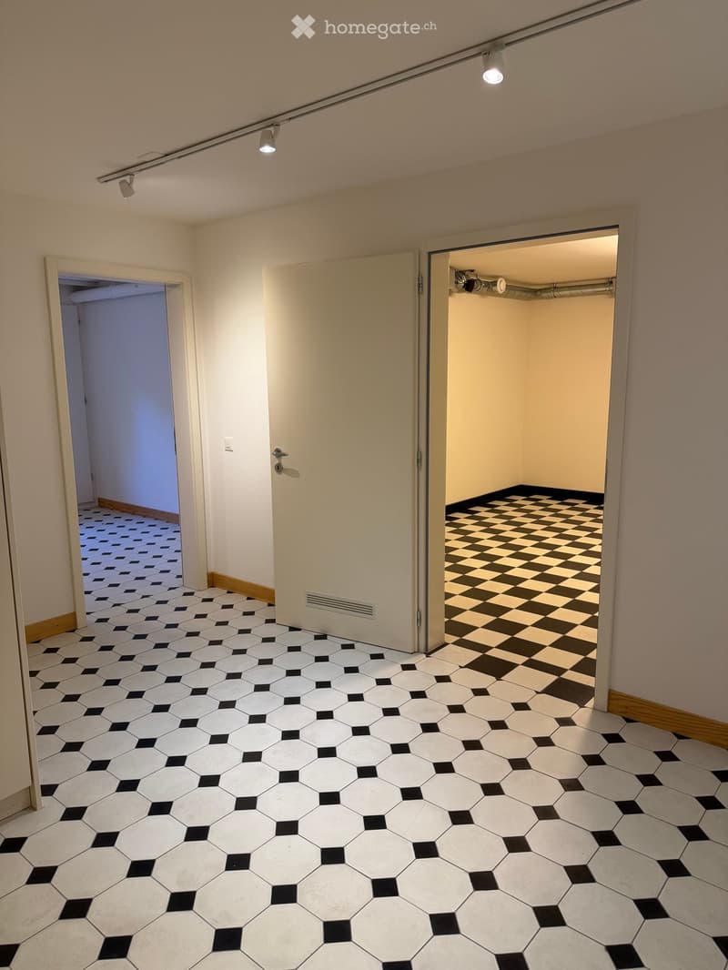 Empty hallway with hexagonal white and black tiled floor, beige walls, and three doors leading to different rooms.