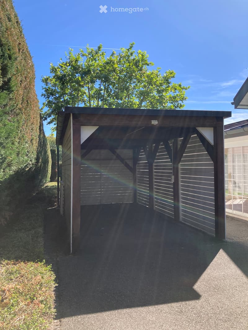 {"type": "Garage", "objects": [], "description": "A modern, wooden garage with a slanted roof and four large, closed garage doors. The surrounding area features a gravel driveway, a lush green hedge on one side, and a tree providing shade.", "localized_de