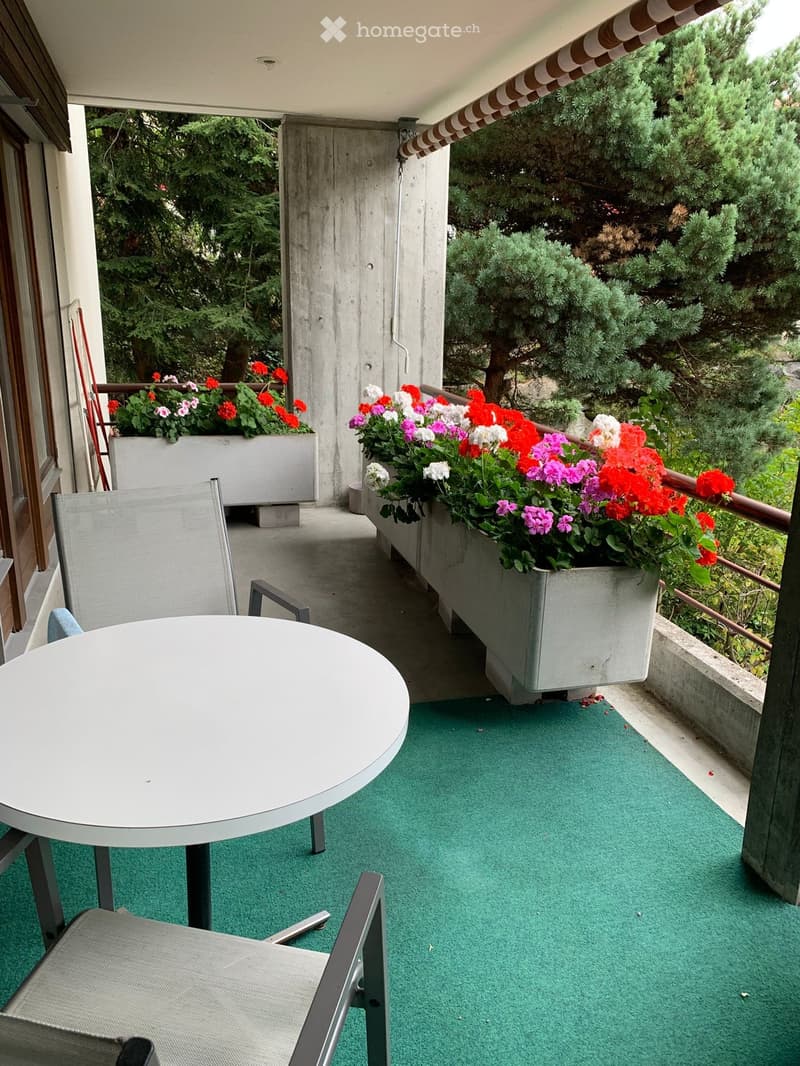 Balcony with green artificial turf, round table, and chair, surrounded by colorful flower boxes and lush greenery.