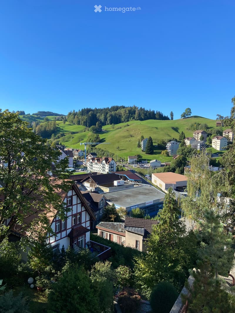 A panoramic view of a town nestled in green hills, with houses, trees, and a clear blue sky.