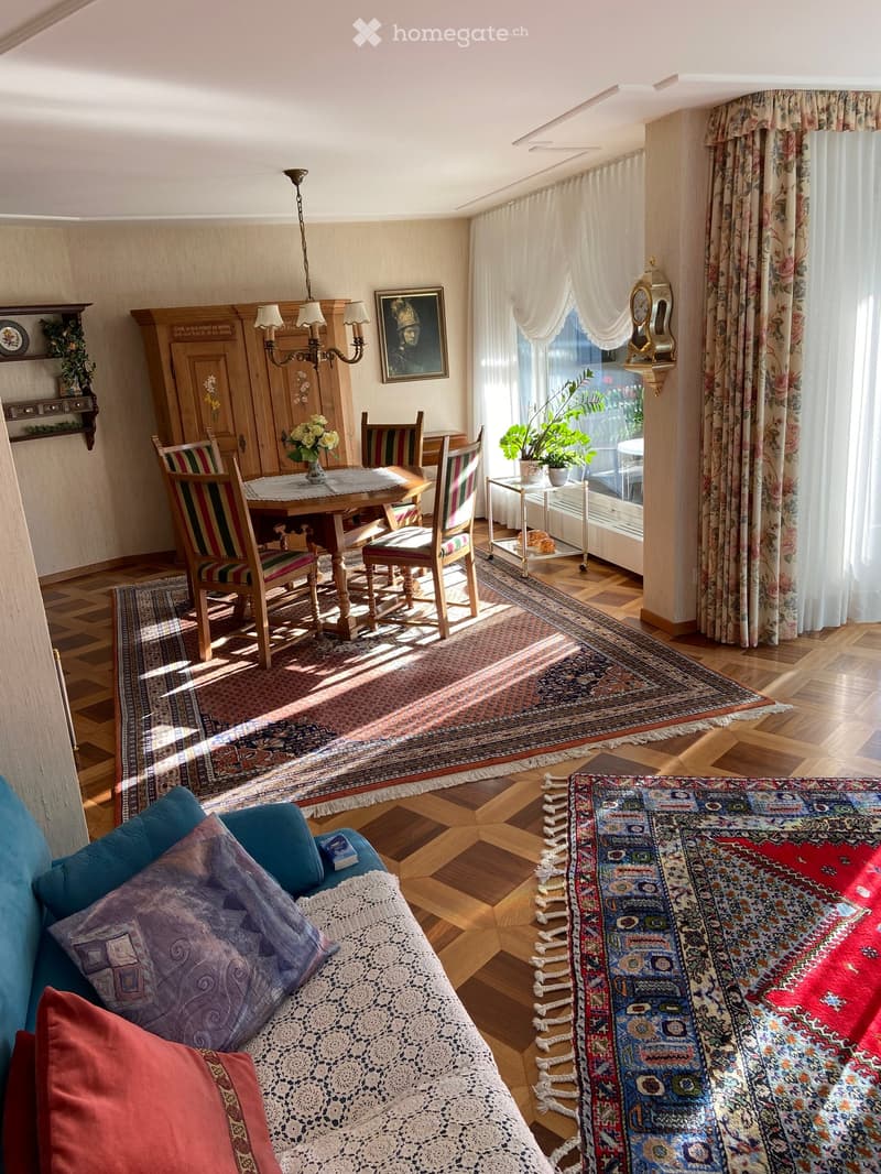 A well-lit living and dining area with wooden floors, a large patterned rug, a sofa with colorful cushions, a dining table with chairs, and a chandelier hanging from the ceiling.