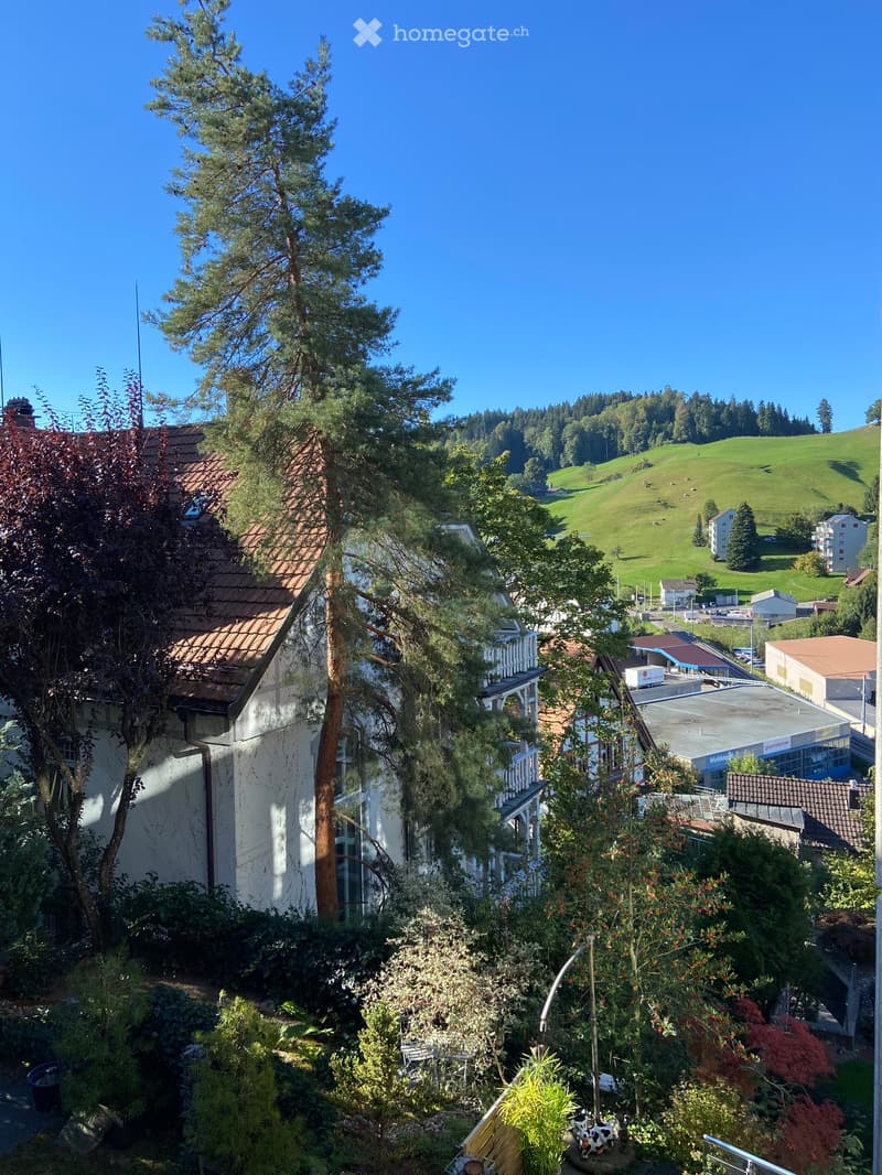 View from a balcony or terrace showing a garden area with various plants and trees, a white building with a tiled roof, and a scenic countryside with green hills and scattered houses in the background.