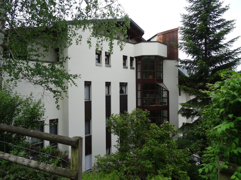 Modern white building with large windows and a balcony, surrounded by greenery and trees.