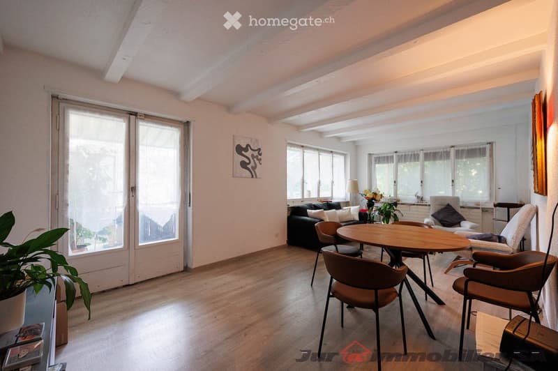 Bright living room with wooden flooring, large windows, and sliding glass doors leading to a balcony. Features a black sofa, wooden dining table with chairs, a floor lamp, and decorative plants.