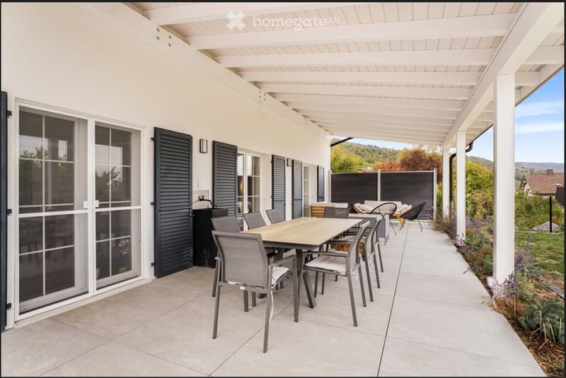 A covered outdoor patio with a wooden table and chairs, large windows, and a scenic view of the surrounding landscape.