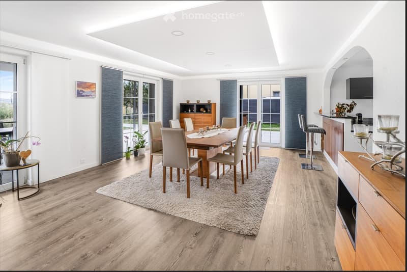 Bright, open dining area with a wooden dining table surrounded by chairs, bar stools near a kitchen island, potted plants, and a side table. Large windows and sliding glass doors lead to an outdoor space.