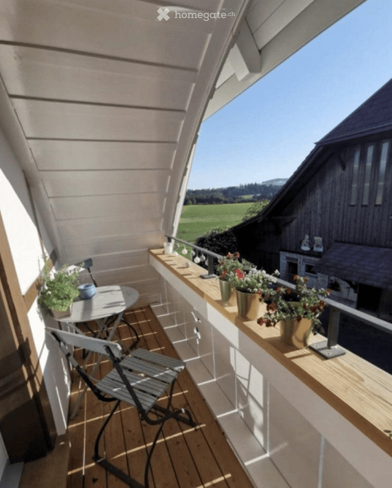 Balcony with wooden decking, small table, chairs, and potted plants, overlooking a green field and mountains.