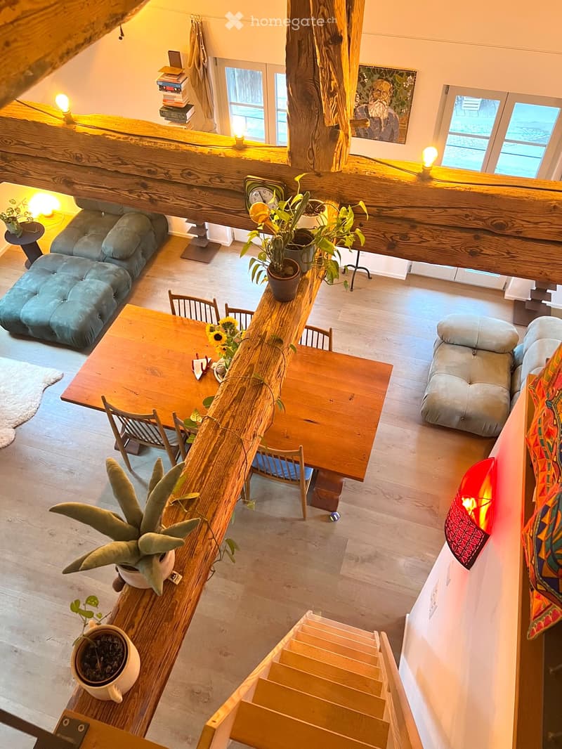 Open-concept living and dining area with wooden floors, large windows, and a staircase leading to an upper level. Features include a sofa, dining table with chairs, potted plants, and decorative lamps.