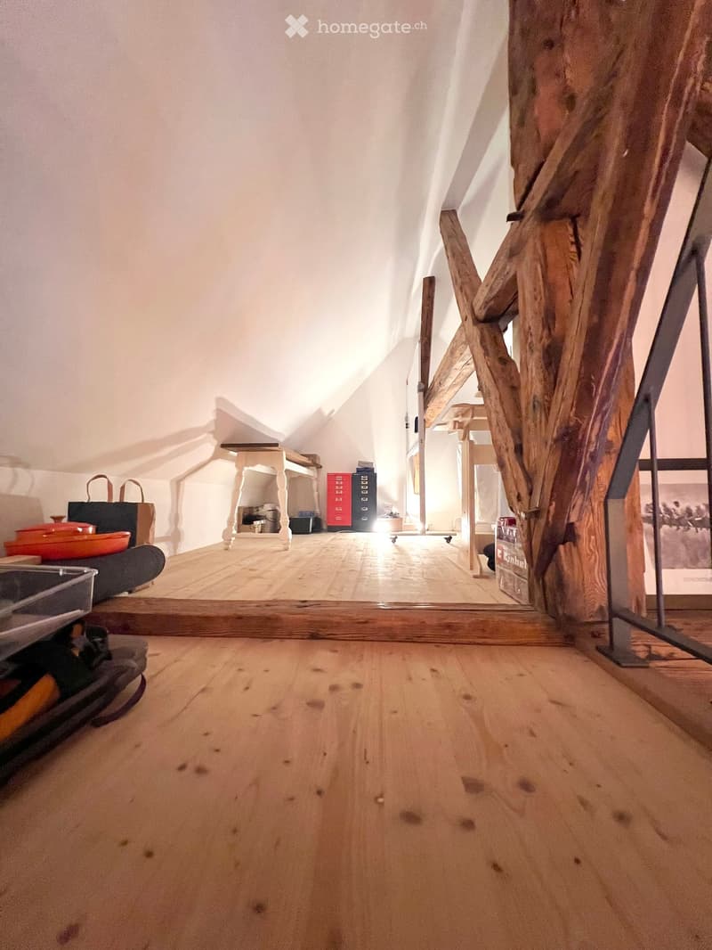 The image shows a wooden floor in a room with a slanted ceiling and exposed wooden beams. There is a small table with a stool, a handbag, and some storage boxes visible. The room appears to be part of an attic or loft space.