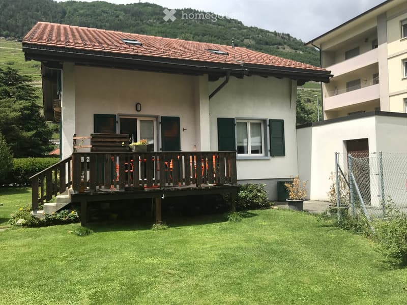 Two-story house with a red-tiled roof, white walls, green shutters, and a wooden balcony with outdoor furniture.