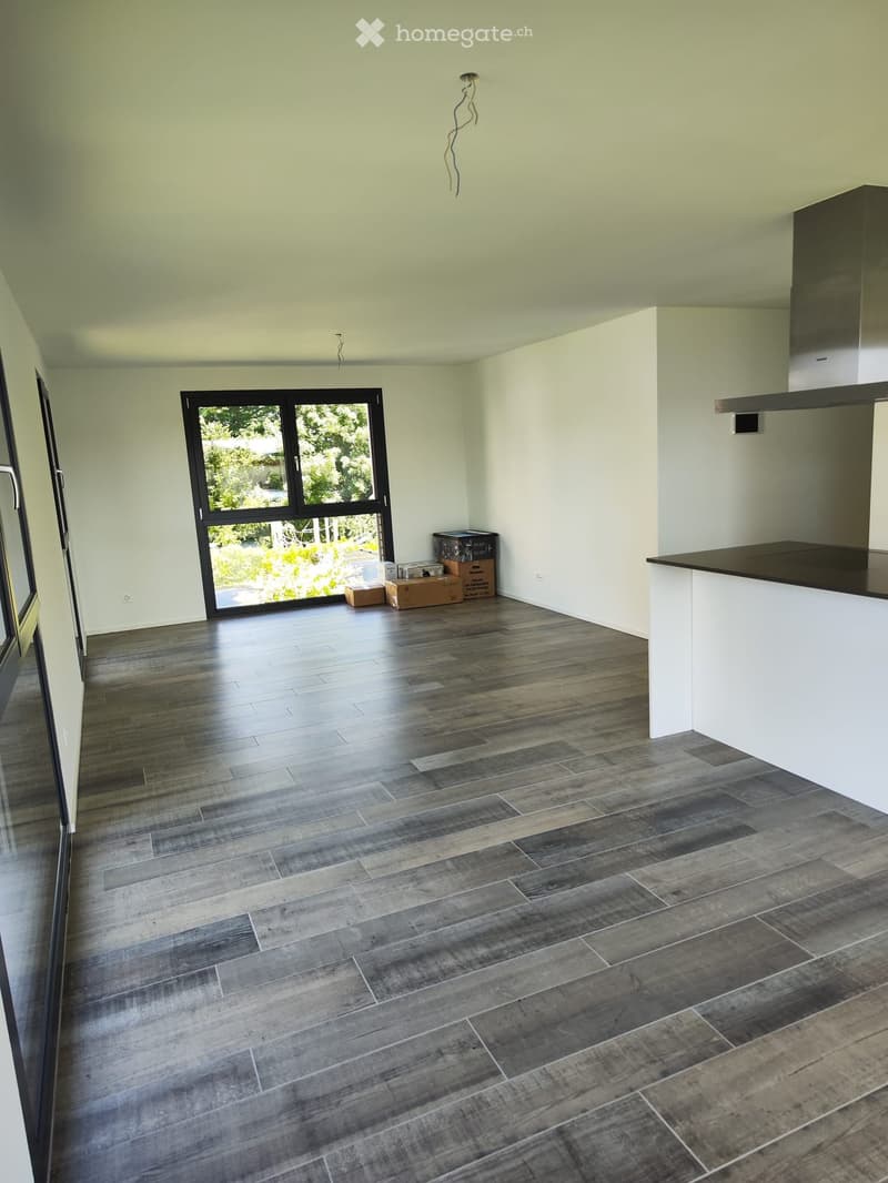 Empty room with light-colored walls, dark wood flooring, and a large window with a view of greenery. There is a small counter area on the right side.