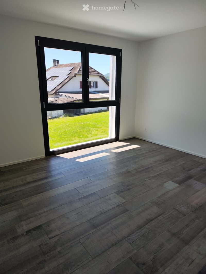 Empty room with wooden flooring and large sliding glass doors leading to a grassy outdoor area and neighboring house with solar panels on the roof.