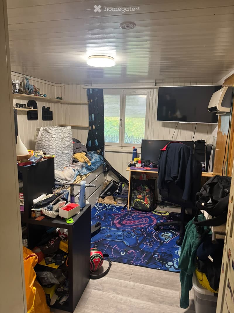 A small, furnished room with a bed, desk, shelves, and a colorful rug. The room features a window with blinds, a TV mounted on the wall, and various personal items scattered around.
