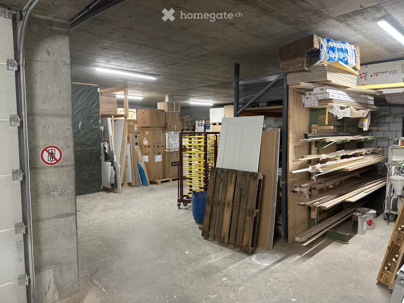 Empty storage area with concrete floors and walls, filled with shelves and stacks of wooden planks, cardboard boxes, and construction materials.